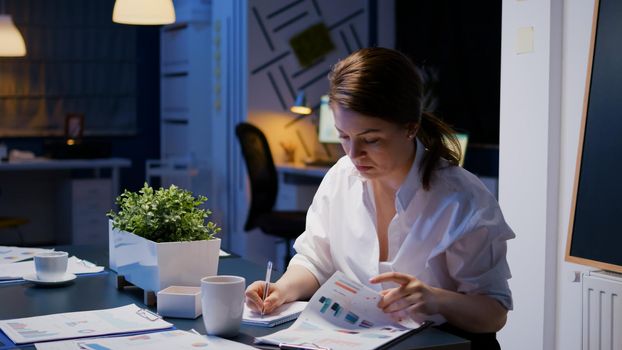 Workaholic businesswoman working in company office meeting room writing financial statistics while looking at reports presentation on monitor late at night. Manager analyzing documents in evening