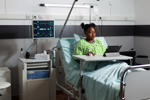 African american patient sitting in hospital ward bed with digital tablet chatting and web surfing. Young person with illness waiting on diagnosis advice while playing with online technology