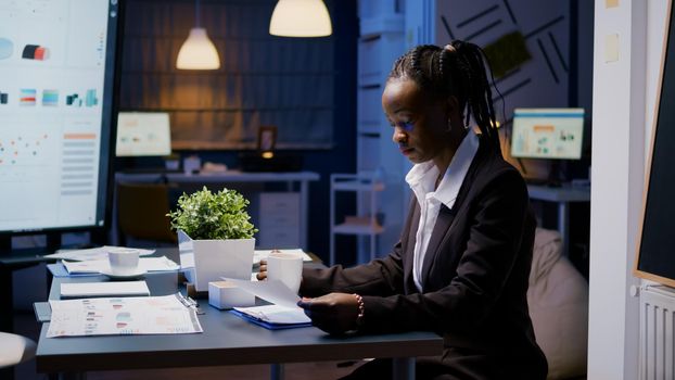 Workaholic african american businesswoman analysing marketing statistics on paperwork drinking coffee sitting at table late at night. Business manager with dark skin working in office meeting room