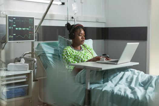 Sick young patient with headphones using laptop in hospital ward. African american teenager with illness, disease recovering in bed while listening to music on online internet technology