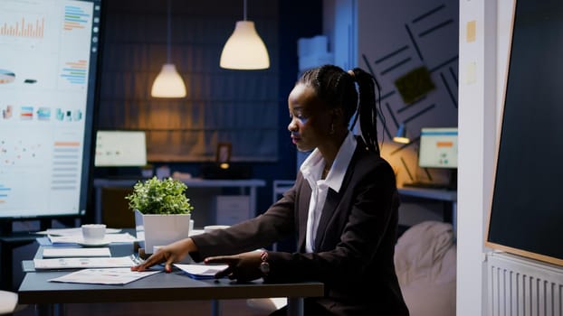 Businesswoman with dark skin hardworking late at night in meeting office room checking company financial charts. Workaholic black manager overworked while drinking coffee analyzing statistics reports