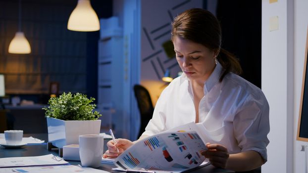 Overworked businesswoman writing financial profit on notebook while working in business company meeting room late at night. Focused workaholic woman analyzing documents statistics charts in evening