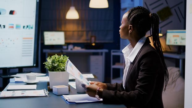 Focused workaholic african american businesswoman working at company financial charts presentation late at night in meeting office room. Overworked manager with dark skin checking profit statistics