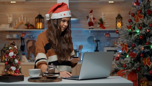 Festive woman holding credit card to shop online for christmas gifts and presents. Young adult with santa hat using laptop in decorated kitchen with seasonal ornaments and tree.