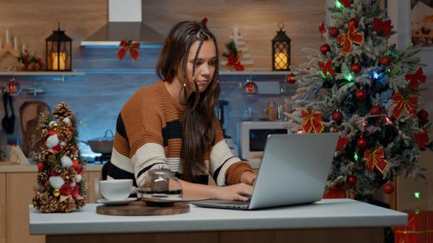 Caucasian woman using laptop in winter decorated kitchen at home. Young adult preparing for christmas eve celebration party waiting for friends checking time for guests to arrive