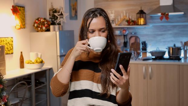 Caucasian adult using video call modern technology on smartphone in kitchen with christmas decorations and ornaments. Woman talking with friends on internet about holiday celebration