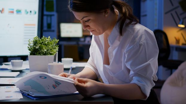 Overworked businesswoman working overtime in business company office meeting room in evening analyzing marketing charts while drinking coffee. Entrepreneur hardworking at financial profit reports