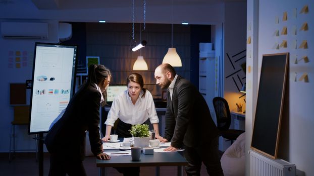 Diverse multi ethnic teamwork standing around table brainstorming company ideas checking management statistics paperwork. Focused coworkers planning overtime business presentation in meeting room