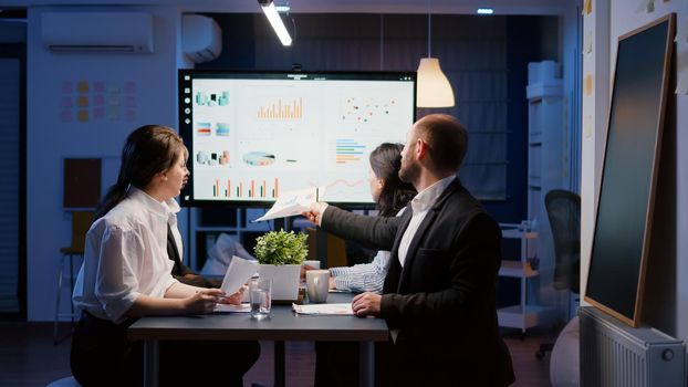 Multi ethnic teamwork of managers analyzing management statistics overworking in office meeting room late at night. Diverse businesspeople looking at financial presentation on monitor sitting at table