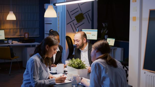 Group of multi-ethnic businesspeople checking company solution analyzing management graphs paperwork brainstorming ideas in office meeting room late at night. Teamwork solving corporate statistics