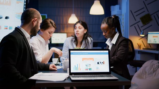 Laptop computer with financial graphs presentation on monitor standing on conference table in meeting room late at night. Diverse multi ethnic teamwork discussing company strategy solving statistics