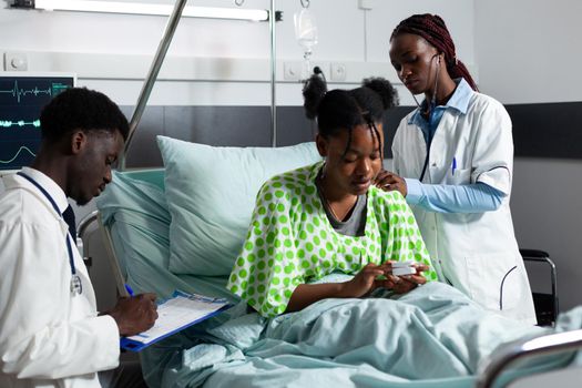 African american woman with doctor occupation consulting patient using stethoscope and medical equipment in hospital ward. Black man writing diagnosis results for young person healthcare