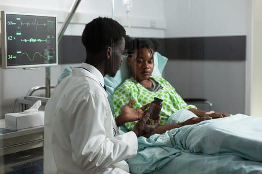 Specialist explaining treatment to ill patient in hospital ward at clinic. African american doctor showing bottle of drugs, pills on prescription for healthcare medicine and immunity