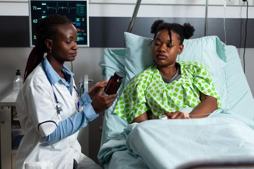 Woman with doctor occupation giving prescription medicine to sick patient laying in hospital ward bed. African american medic holding bottle of pills, drugs, treatment for disease