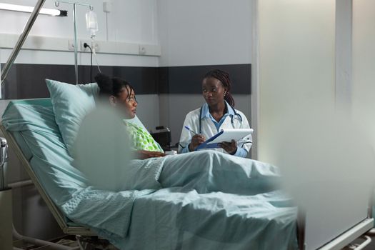 Doctor of african ethnicity giving disease advice to teenager sitting in hospital ward bed at clinic. African american medic woman helping young woman, patient with sickness treatment