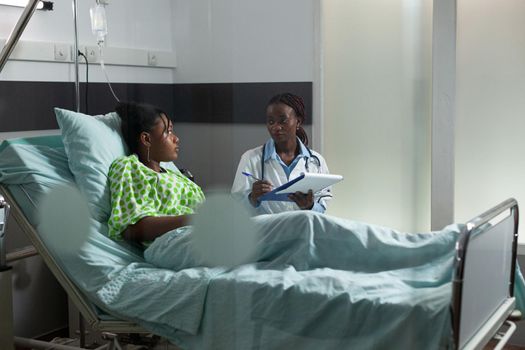 African american young adult with disease talking to doctor while sitting in hospital ward. Black surgeon listening to ill patient in bed, helping with professional advice and medicine