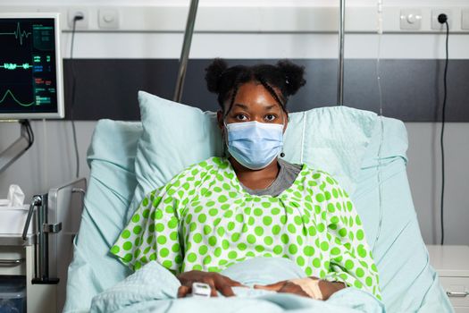 Portrait of african american patient with face mask looking at camera sitting in hospital ward bed. Young woman with disease, illness waiting on consultation with medical equipment