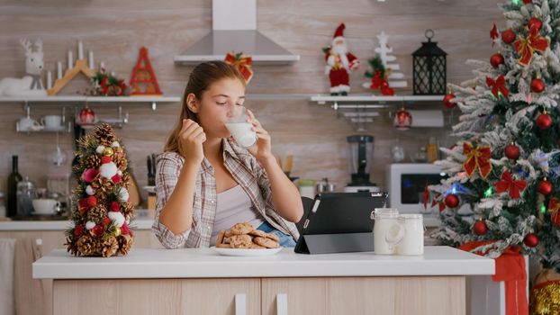 Portrait of children eating baked delicious cookies drinking milk while watching online xmas video on tablet computer in decorated kitchen. Caucasian girl celebrating christmas enjoying winter holiday