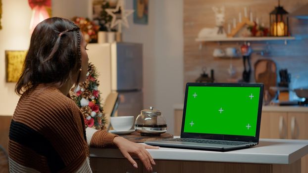 Young adult looking at green screen on laptop in christmas decorated kitchen. Caucasian woman using digital technology device waiting for seasonal festive celebration dinner and friends