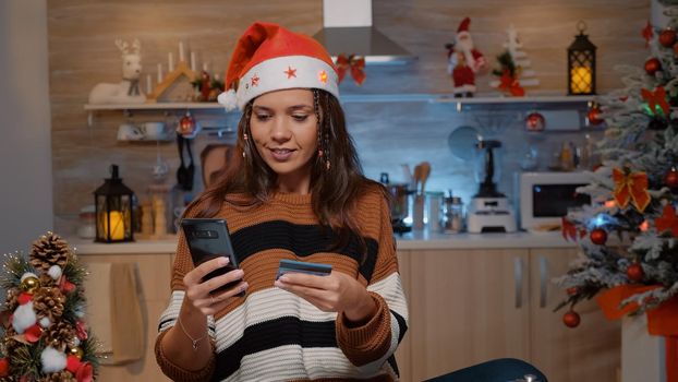 Festive woman using smartphone for shopping presents with credit card in winter decorated kitchen. Young adult getting gifts for relatives at christmas eve dinner celebration festivity