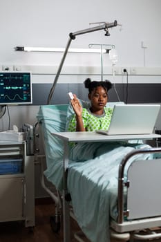 African american girl in hospital ward using video call technology on laptop. Sick young person sitting in bed with modern device for communication and conference on internet connection