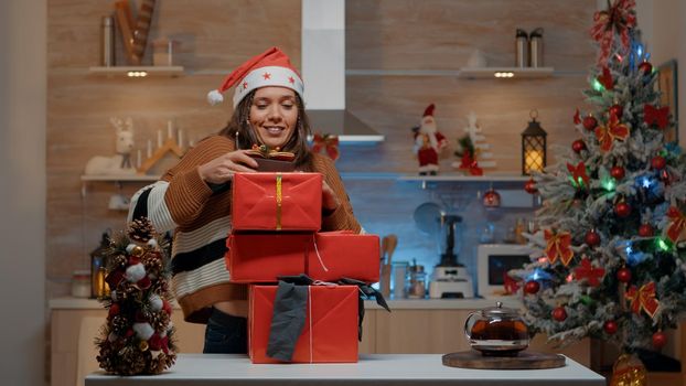 Woman with santa hat carrying gifts in decorated kitchen for christmas eve celebration party. Young adult holding presents to give to friends and family and seasonal festivity