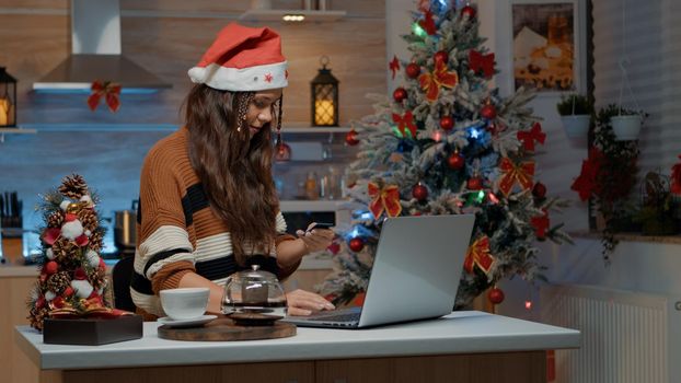 Young woman doing online shopping on laptop using credit card to buy presents for friends and family. Caucasian adult preparing for christmas eve celebration dinner with festive gifts