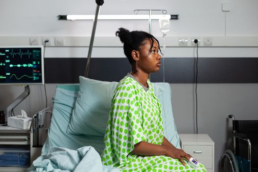 Sick patient of african american ethnicity laying on bed in hospital ward. Black teenager sitting with heart rate monitor, oximeter and IV drip bag at healthcare recovery clinic for treatment