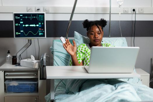 African american girl in hospital ward using video call technology on laptop. Sick young person sitting in bed with modern device for communication and conference on internet connection