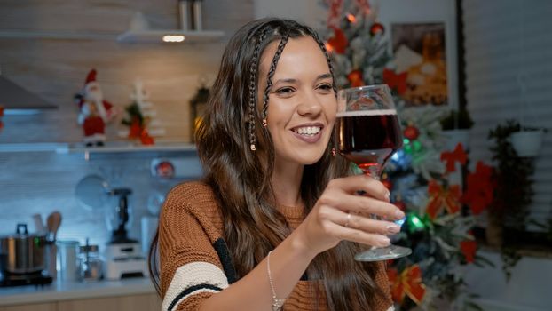 Happy young woman talking on video call at home in festive kitchen with christmas decorations and ornaments. Adult preparing to bake for seasonal dinner party with friends and family