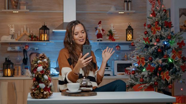 Cheerful woman on christmas video call with friends at home in winter decorated kitchen. Young adult celebrating festivity with ornaments and decorations for seasonal holiday dinner