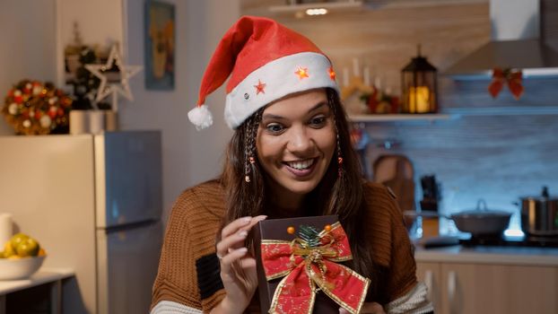Young festive woman showing gifts to friends while using video call communication in kitchen with seasonal ornaments and decorations. Person preparing for christmas eve dinner party
