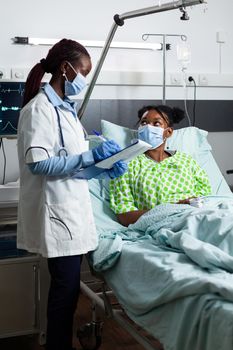 Black doctor talking to african american young patient laying in hospital ward bed. Woman checking healthcare diagnosis while girl sitting with medical equipment and technology for recovery