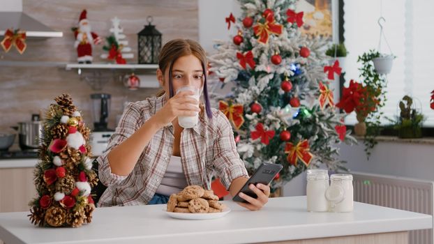 Portrait of children sitting at table in xmas decorated kitchen browsing on social media using smartphone. Caucasian girl enjoying christmas holiday eating traditional homemade cookies drinking milk