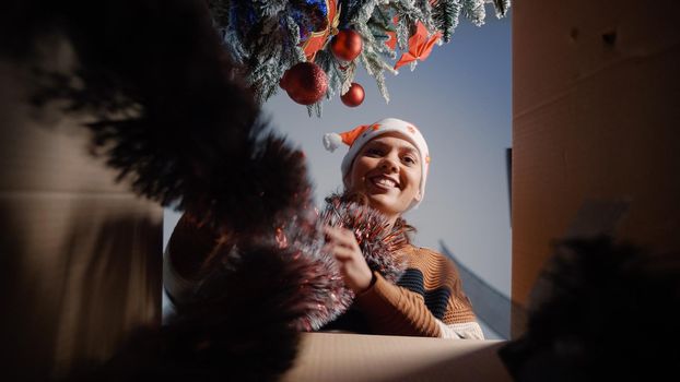 Woman opening box of christmas ornaments for winter season decorating at home. Young caucasian adult getting excited about garlands and globes for green tree preparing for festivity