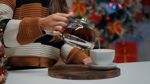 Close up of woman pouring tea from kettle in cup on kitchen counter at christmas prepared home. Person getting a hot drink in mug for cozy atmosphere while waiting on holiday celebration