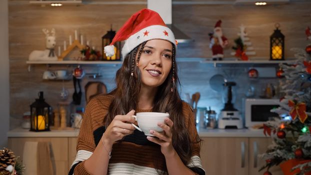 Happy woman with santa hat thinking about christmas time while holding cup of tea in hands at festive designed home. Young person preparing for holiday dinner party and gifts for family