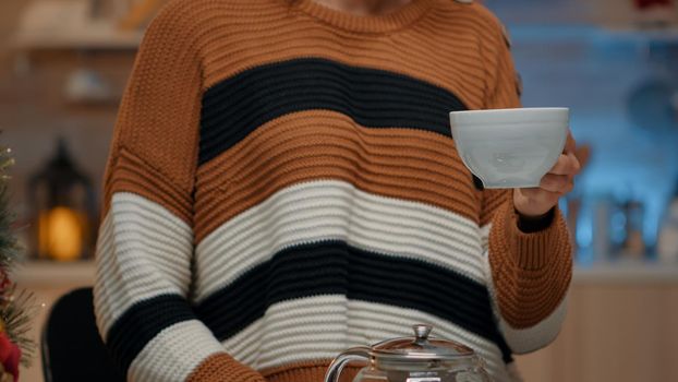 Close up of young adult holding cup of tea in kitchen with utensils designed for festive christmas eve celebration party at home. Woman preparing winter beverage from kettle on counter
