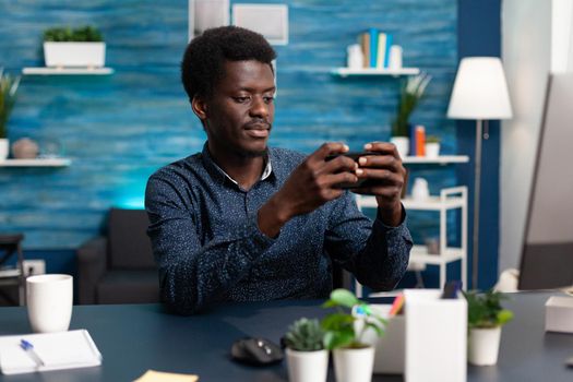 African american man smiling while using a smartphone at desk to check social media. Remote working guy taking a break from assignment, using phone for relaxation and communication