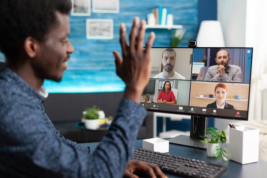 Black man waving to colleagues on video call chat using online internet and webcam communication. Remote worker working from home keeping distance while using technology and computer