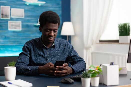 Black african american man using a smartphone at home working on social media platform. Young guy looking at telephone sitting at desk doing online internet search on modern device