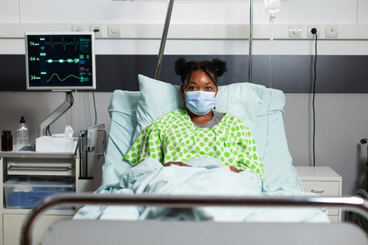Portrait of young patient with disease sitting in hospital ward at healthcare clinic. African american sick person with face mask and oximeter laying in bed with medical equipment