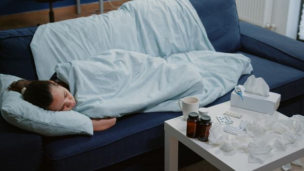 Person with disease sleeping wrapped in blanket on sofa while feeling cold and shivering. Woman with sickness laying at home trying to cure virus infections and flu with fever.