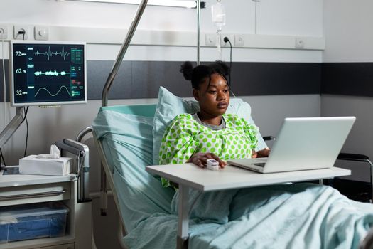 Young woman of african ethnicity browsing internet on laptop in hospital ward bed. African american adult with sickness, disease web surfing on digital device and computer technology