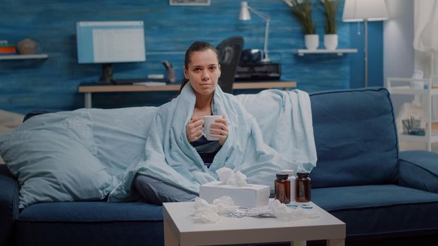 Portrait of woman with flu holding cup of tea in blanket while looking at camera. Sick person with coronavirus symptoms having medicaments, pills, drugs, capsules and treatment for healing