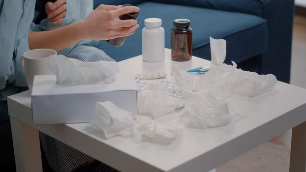 Close up of hand searching for medical treatment against flu, reading label of tablets with capsules and bottle with pills. Table with medication, tissues, cup of tea and thermometer