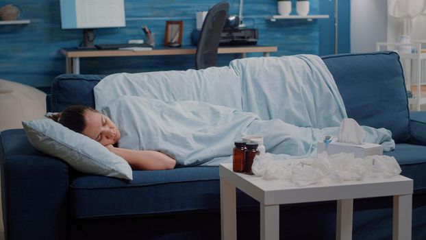 Ill person with sickness sleeping wrapped in blanket. Sick woman with virus disease resting on couch while having medicaments, bottles of pills, tissues, thermometer, tea on table.