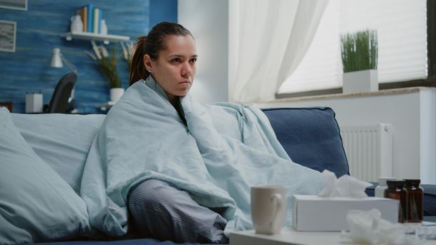 Sick woman with seasonal flu shivering in blanket at home. Cold adult sitting in living room with medicaments, treatment and cup of tea to cure virus infection and disease. Ill person