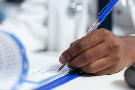 Closeup of african american doctor hand analyzing ill symptoms writing medical treatment on clipboard. Therapist man working at medicine prescription for sick patient in hospital office