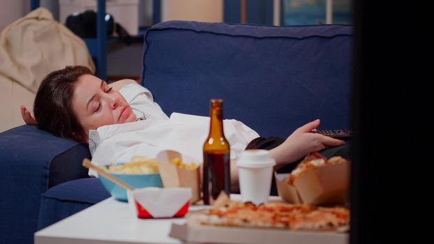 Young adult feeling sleepy while watching television on sofa in living room. Tired woman falling asleep with remote control after work and eating fast food meal, laying on couch at home